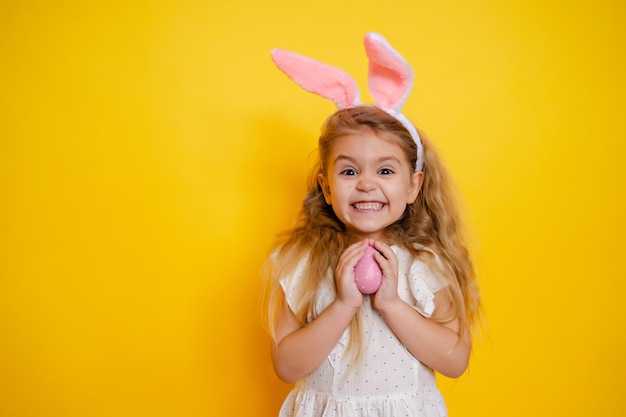 Ragazza bionda sorridente impertinente con le orecchie del coniglietto che tiene un uovo di Pasqua nelle sue mani