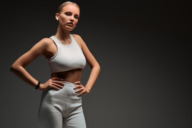 Ragazza bionda sportiva in abiti da allenamento bianchi con le cuffie in studio su sfondo grigio