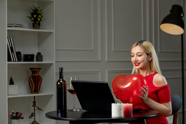 Ragazza bionda sveglia di amore virtuale in vestito rosso alla data di distanza con il palloncino del cuore della holding del vino