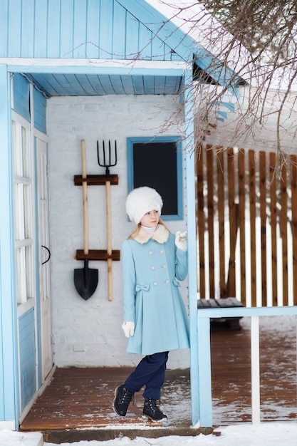 Ragazza bionda sveglia in un cappotto blu e cappello di pelliccia bianco in inverno. Tempo nevoso. La ragazza sotto il portico della casa. Casa invernale. Modello in posa sulla strada. Il concetto di vacanze invernali.