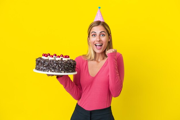 Ragazza bionda uruguaiana con torta di compleanno isolata su sfondo giallo che celebra una vittoria in posizione di vincitore