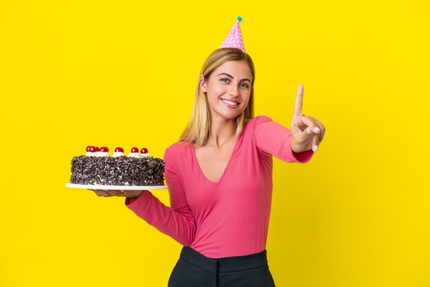 Ragazza bionda uruguaiana con torta di compleanno isolata su sfondo giallo che mostra e solleva un dito
