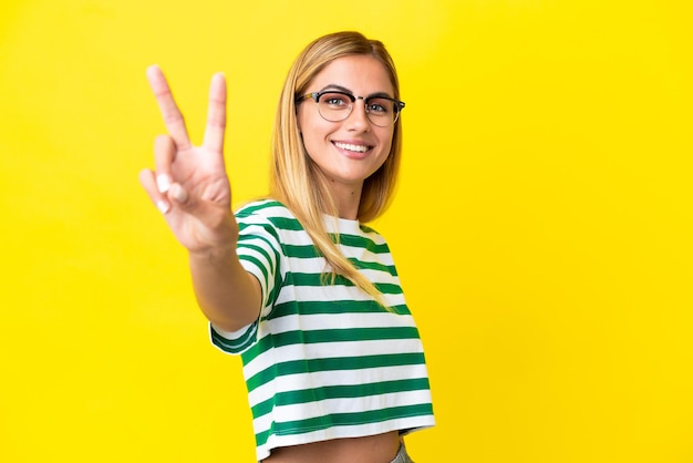 Ragazza bionda uruguaiana isolata su sfondo giallo sorridente e che mostra il segno della vittoria