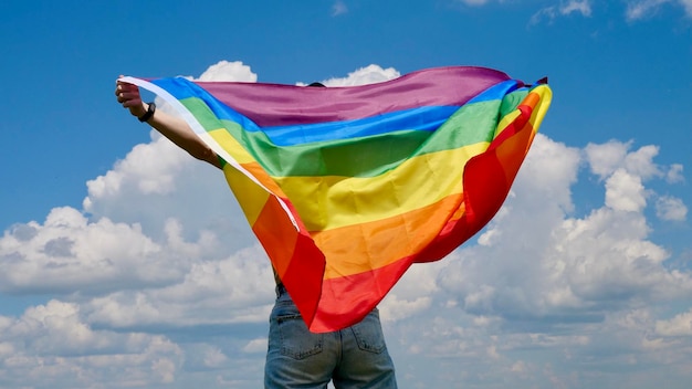 Ragazza bisessuale donna lesbica transgender omosessuale che tiene in mano una bandiera dell'identità di genere LGBT arcobaleno sullo sfondo del cielo con nuvole in una giornata di sole e celebra una parata gay nel mese dell'orgoglio