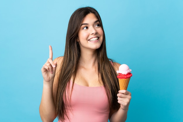Ragazza brasiliana dell'adolescente che tiene un gelato della cornetta sopra fondo blu isolato che intende realizzare la soluzione mentre sollevando un dito su