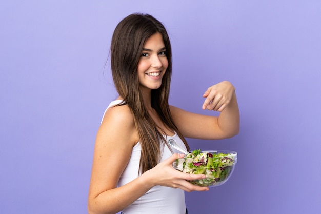 Ragazza brasiliana dell'adolescente che tiene un'insalata sopra fondo porpora isolato