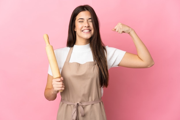 Ragazza brasiliana dell'adolescente che tiene un mattarello isolato sulla parete rosa che fa forte gesto