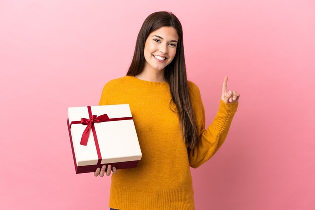 Ragazza brasiliana dell'adolescente che tiene un regalo sopra fondo rosa isolato che indica su una grande idea