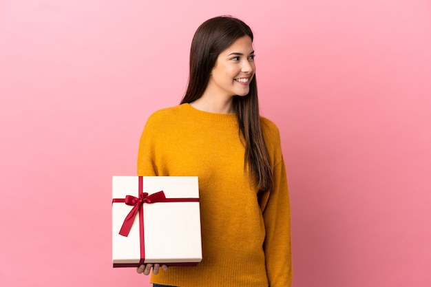 Ragazza brasiliana dell'adolescente che tiene un regalo sopra fondo rosa isolato che osserva al lato