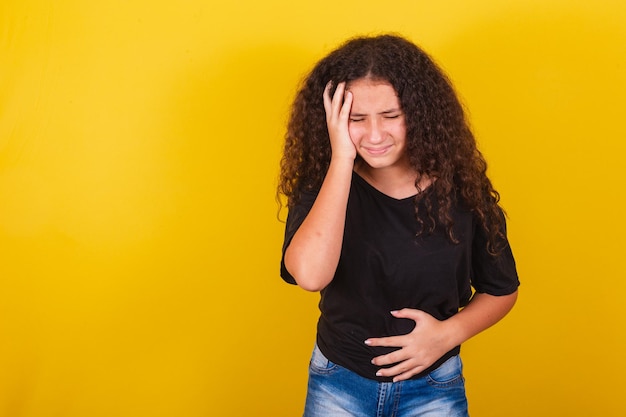 Ragazza brasiliana latinoamericana per capelli afro sfondo giallo Mani sulla pancia e sulla fronte mal di pancia disagio addominale coliche diarrea mal di testa stanchezza