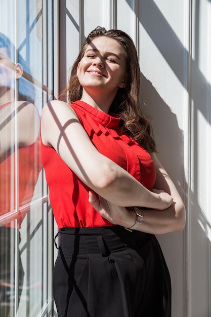 Ragazza bruna alla moda in una camicetta rossa posa vicino alla finestra e gode della luce del sole e del calore.