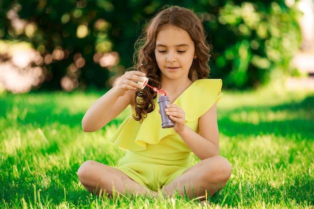 Ragazza bruna con i capelli scuri si siede sull'erba nel parco e soffia bolle. ritratto di bambini. infanzia felice. Foto di alta qualità