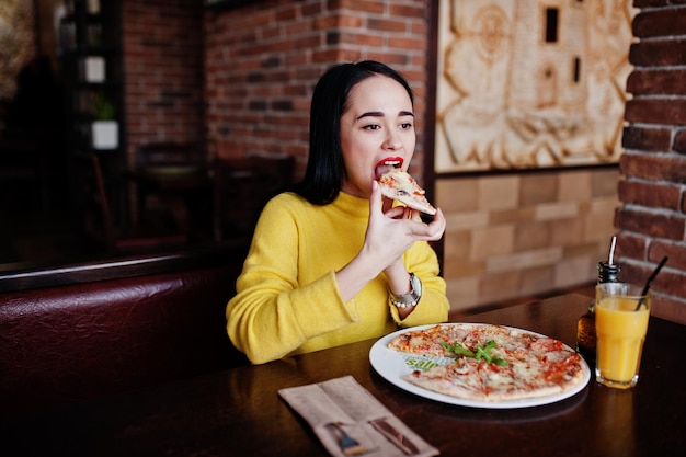 Ragazza bruna divertente in maglione giallo che mangia pizza al ristorante