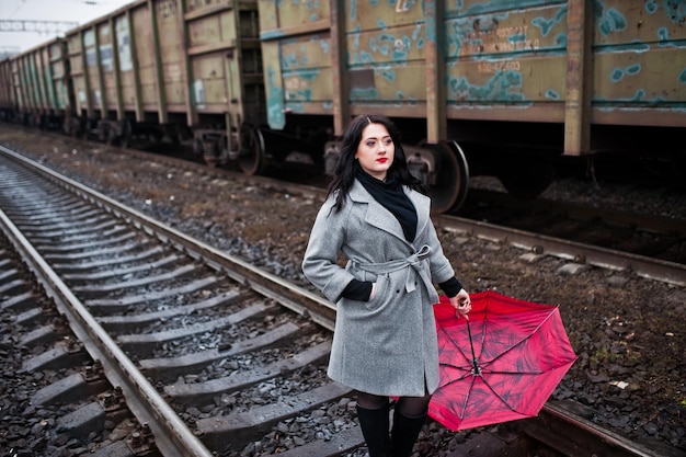 Ragazza bruna in cappotto grigio con ombrello rosso nella stazione ferroviaria