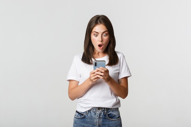 Ragazza bruna scioccata e colpita guardando stupito lo schermo dello smartphone.