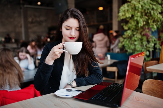 Ragazza bruna seduta sul caffè con una tazza di cappuccino, lavorando con il portatile rosso.