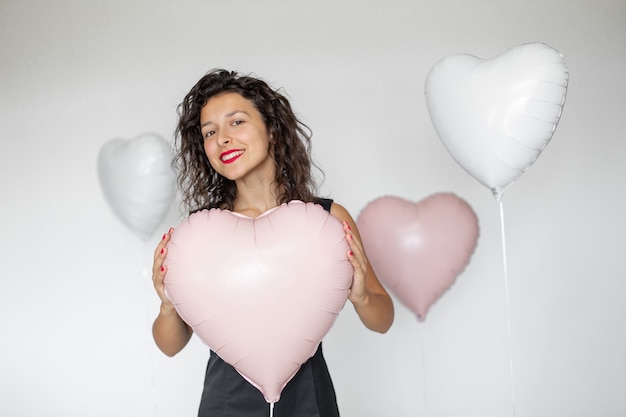 Ragazza bruna sexy in posa con palloncini a forma di cuore su sfondo bianco.