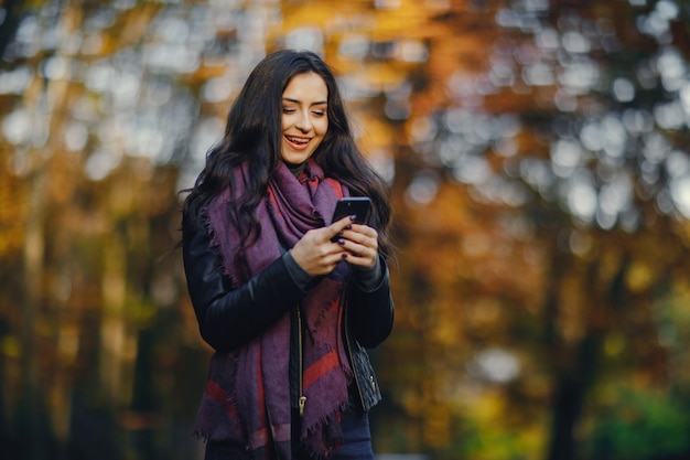 ragazza bruna usando il suo telefono mentre si sta rilassando al parco durante l&#39;autunno