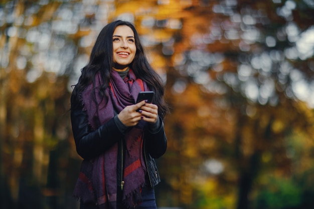 ragazza bruna usando il suo telefono mentre si sta rilassando al parco durante l&#39;autunno