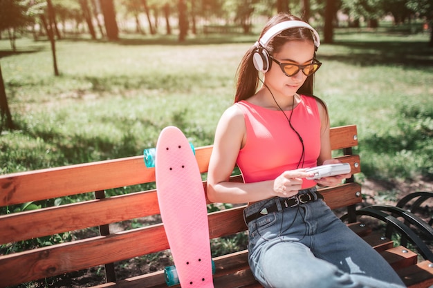 Ragazza calma e pacifica è seduto sulla panchina fuori e guardando il suo lettore musicale. Indossa occhiali divertenti.