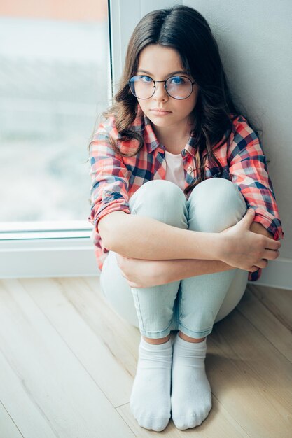 Ragazza calma in jeans e camicia a scacchi che le abbraccia le gambe mentre è seduta vicino al muro vicino alla finestra