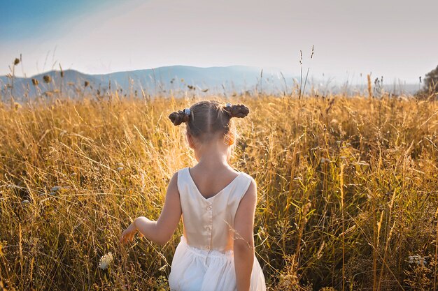 Ragazza carina che balla attraverso un bellissimo prato con grano e fiori in montagna, vista posteriore