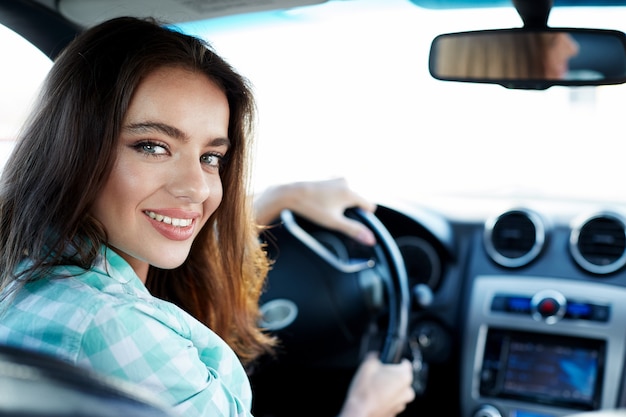 Ragazza carina che indossa la camicia blu seduto in una nuova automobile, bloccato nel traffico, ritratto, acquisto di auto nuove, autista donna, sorridente e guardando la fotocamera.