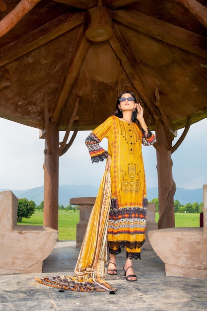 Ragazza carina che indossa un vestito giallo completo e occhiali per un servizio fotografico di moda sotto una capanna in giardino