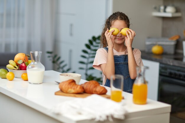 Ragazza carina che tiene dei limoni vicino agli occhi su sfondo sfocato della cucina, vicino al tavolo.