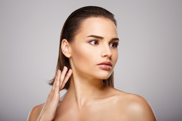 Ragazza carina con capelli castani, pelle fresca e pulita, occhi grandi e spalle nude che tengono la mano vicino al viso in posa al muro grigio, un modello con trucco nudo chiaro.