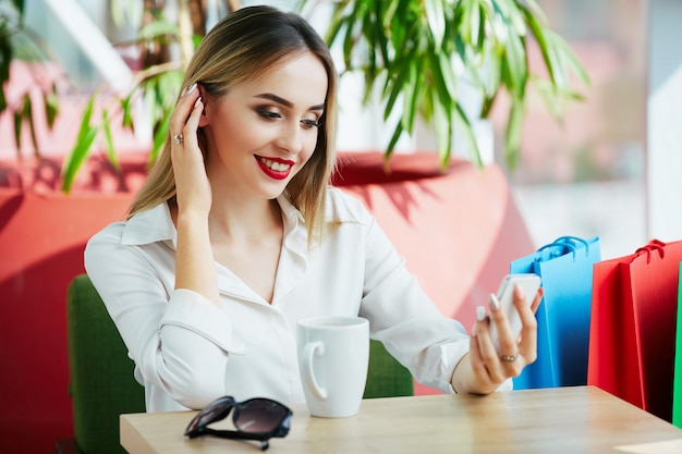 Ragazza carina con capelli castano chiaro e labbra rosse che indossa camicetta bianca e seduta con borse della spesa colorate e tazza di caffè, tenendo il telefono cellulare, concetto di acquisto.