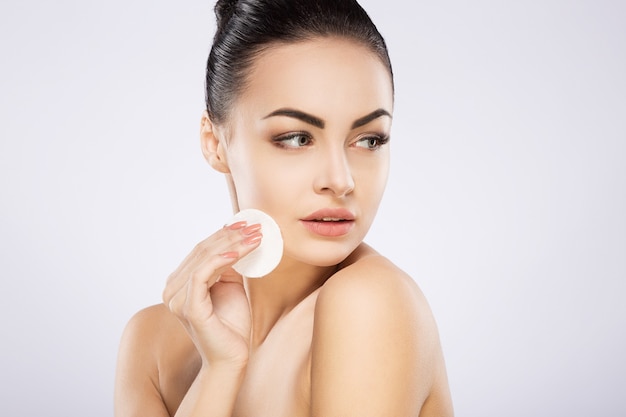 Ragazza carina con capelli neri fissati dietro, grandi occhi, sopracciglia folte e spalle nude in piedi su sfondo grigio studio e che tiene la spugna per la pulizia cosmetica, ritratto.