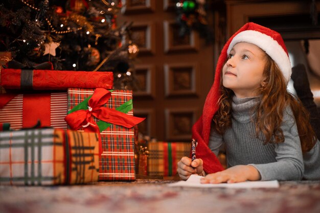 Ragazza carina con cappello da Babbo Natale che scrive una lettera a Babbo Natale nel soggiorno decorato di Natale vista ravvicinata