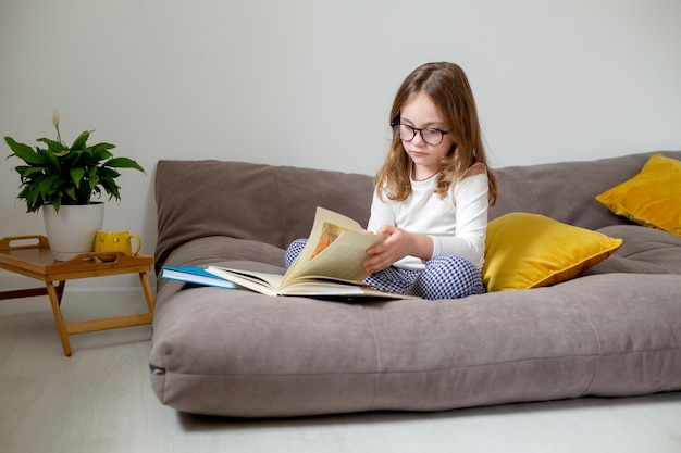 ragazza carina con gli occhiali jeans e un dolcevita bianco sta leggendo un libro seduta sul letto