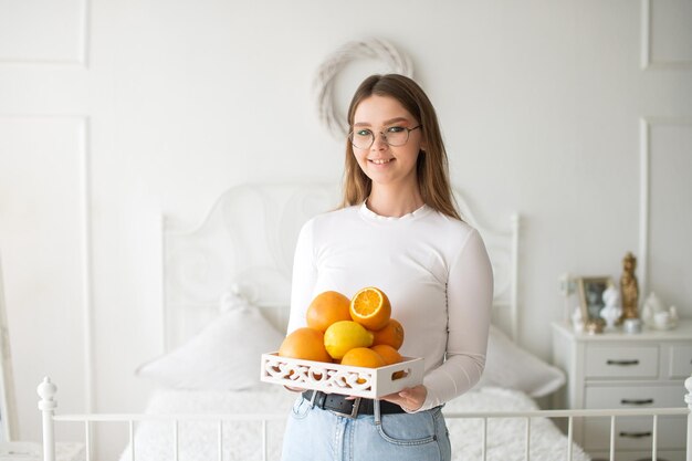 Ragazza carina con gli occhiali tiene un cesto di frutta e sorride in una stanza bianca