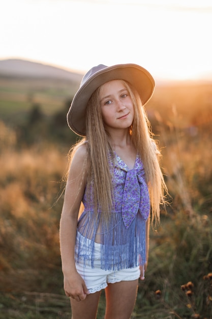 Ragazza carina con lunghi capelli biondi, indossa cappello marrone, in posa nel campo