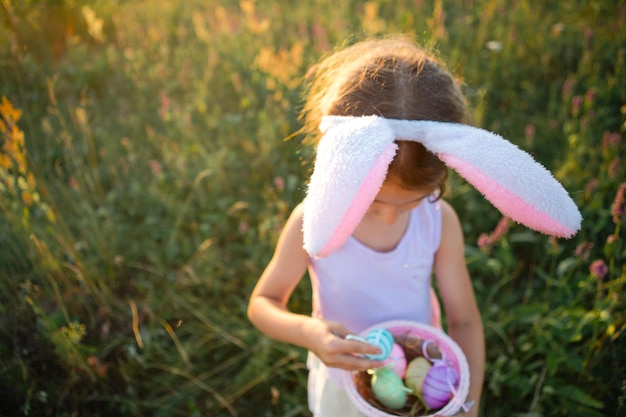 Ragazza carina e divertente con uova di Pasqua dipinte in primavera in natura in un campo con luce solare dorata e fiori. Vacanze di Pasqua, coniglietto di Pasqua con le orecchie, uova colorate in un cestino. Stile di vita