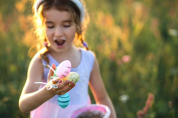 Ragazza carina e divertente con uova di Pasqua dipinte in primavera in natura in un campo con luce solare dorata e fiori. Vacanze di Pasqua, coniglietto di Pasqua con le orecchie, uova colorate in un cestino. Stile di vita