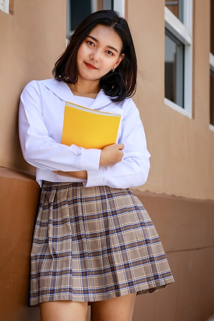 Ragazza carina e giovane che indossa l'uniforme da scolaretta in stile giapponese e coreano che tiene il libro e posa davanti alla telecamera con divertimento e felicità di fronte all'edificio scolastico.