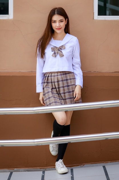 Ragazza carina e giovane che indossa l'uniforme da scolaretta in stile giapponese e coreano e posa davanti alla telecamera con divertimento e felicità di fronte all'edificio scolastico.