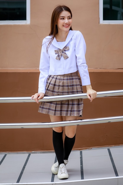 Ragazza carina e giovane che indossa l'uniforme da scolaretta in stile giapponese e coreano e posa davanti alla telecamera con divertimento e felicità di fronte all'edificio scolastico.