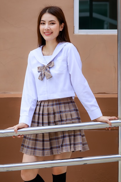 Ragazza carina e giovane che indossa l'uniforme da scolaretta in stile giapponese e coreano e posa davanti alla telecamera con divertimento e felicità di fronte all'edificio scolastico.