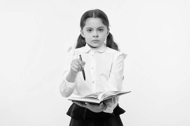 Ragazza carina faccia seria sfondo giallo Bambino ragazza uniforme scolastica vestiti tiene libro e penna Controlla i miei compiti Uniforme scolastica bambino bambino intelligente fatto con i compiti Controlla il concetto di conoscenza