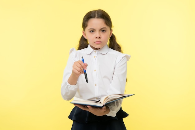 Ragazza carina faccia seria sfondo giallo. I vestiti dell'uniforme scolastica della ragazza del bambino tengono il libro e la penna. Controlla i miei compiti. Bambino intelligente uniforme scolastica bambino fatto con i compiti. Controllare il concetto di conoscenza.