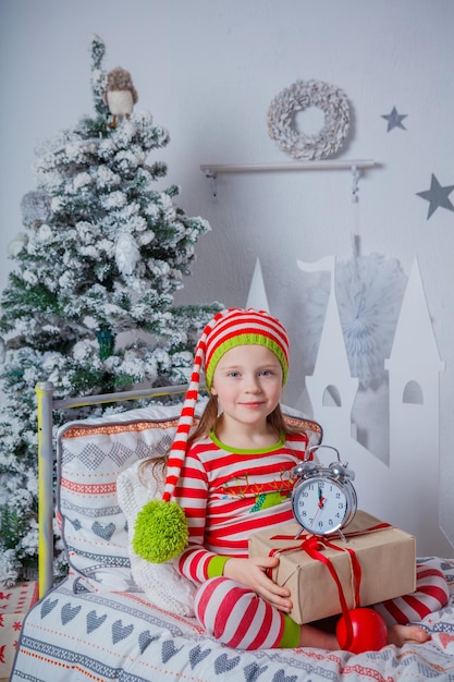 Ragazza carina felice sorridente allegra vestita in pigiama a righe con regalo e orologio seduto nella stanza decorata di Capodanno a casa. Buon umore natalizio. Stile di vita, famiglia e concetto di vacanza 2018