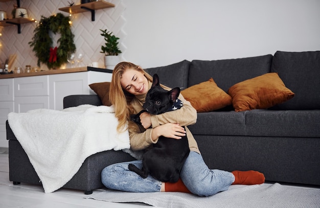 Ragazza carina in abiti casual seduta vicino al divano in casa e abbracciando il cane.