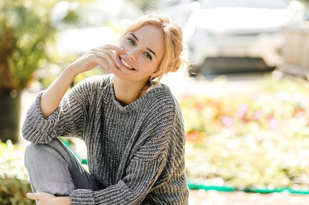 Ragazza carina in maglione lavorato a maglia e jeans grigi seduto su una panchina in giardino e sorridente