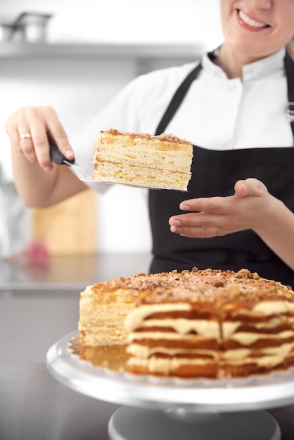 Ragazza carina pasticcere in uniforme dimostra una torta nelle sue mani nel telaio verticale della cucina