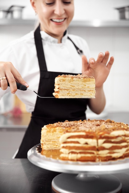 Ragazza carina pasticcere in uniforme dimostra una torta nelle sue mani nel telaio verticale della cucina