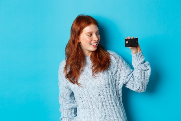 Ragazza carina rossa in maglione che mostra la carta di credito, sorride alla macchina fotografica, in piedi su sfondo blu